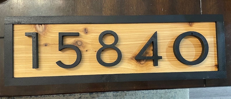 May include: Wooden address sign featuring the numbers 15840 in black against a light brown wood grain background. The numbers are raised and have a modern font. The sign is framed with a black border.
