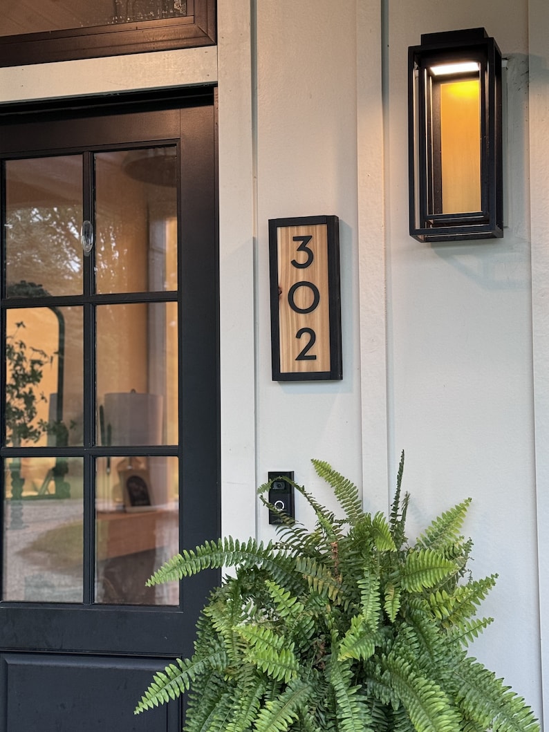 May include: Wooden house number sign displaying the number "302" in a black font. The sign is framed in black and mounted on a light-colored wall. A black outdoor light fixture and a fern plant are also visible.