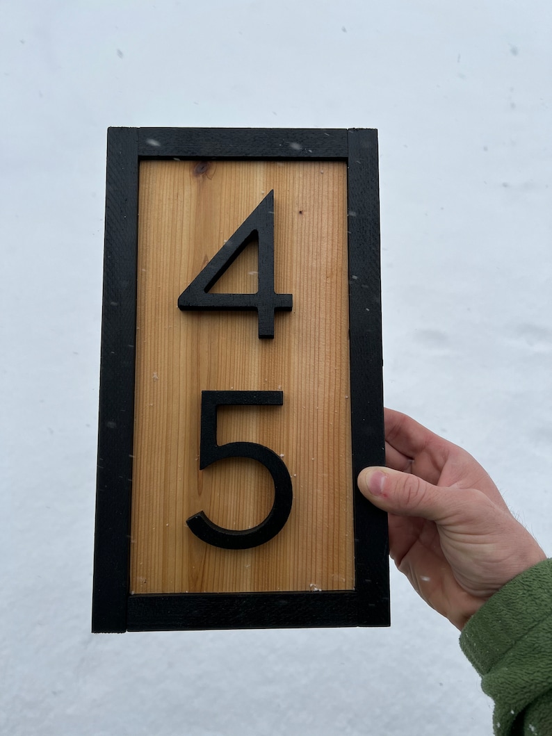 May include: Wooden address sign with the numbers 4 and 5 in black. The numbers are mounted on a light brown wooden plank, framed by a black border. The sign is held against a snowy background.