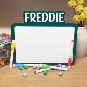 May include: A green framed whiteboard with the name "FREDDIE" in white letters at the top. Several colorful markers and decorative balls are scattered in front of the board. A vase of yellow flowers is in the background.
