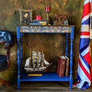 May include: A blue side table with a decorative floral pattern on the top shelf. The table has two shelves and a ship model on the lower shelf. There are books and a candle on the top shelf. A framed portrait of a man in a black suit and a lion figurine are also on the top shelf.