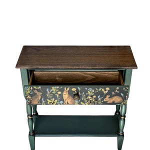Top view of a hand-painted green console table with an open drawer, showing the woodland rabbit motif on the front panel. The piece features a dark wood top, turned legs, gold accents, and a lower shelf, shown on a white background.