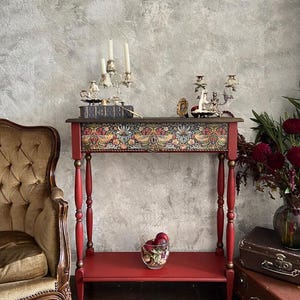 May include: A red wooden console table with a single shelf and decorative turned legs. The table top features a floral patterned design with birds. A book, candlesticks, and a small bowl of red berries are on the table.