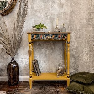 Puede incluir: Una mesa auxiliar pintada de amarillo con un cajón y un estante inferior. El frente del cajón presenta una impresión colorida de pájaros y follaje. La mesa tiene patas torneadas y se encuentra sobre un suelo de madera oscura.