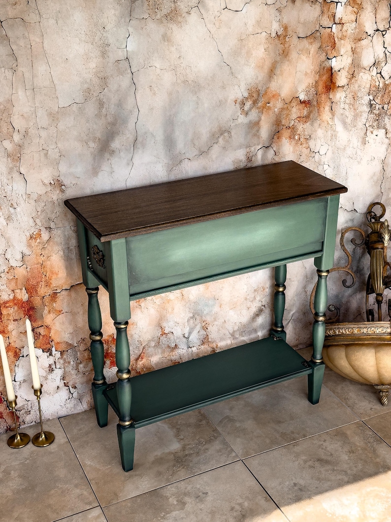 May include: A green and brown console table with a dark wood top and a lower shelf. The table has decorative gold accents on the legs and trim. The table is approximately 36 inches long, 12 inches wide, and 30 inches tall.