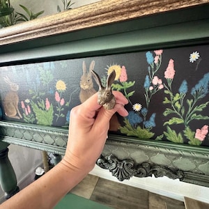 May include: A bronze-colored rabbit figurine is held in front of a dark green and black painted surface. The surface features a floral and rabbit design. The figurine is a decorative accent piece. The background includes a wooden frame and green painted furniture.