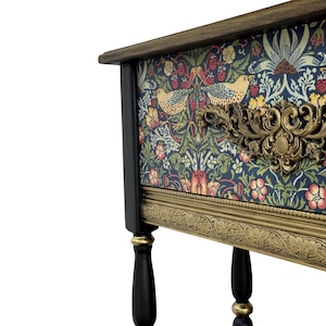 May include: A small, decorative cabinet with a dark wood frame and black legs. The cabinet features a floral patterned fabric on the front and sides, with a brass-colored ornate handle and trim. The cabinet's dimensions are not visible.