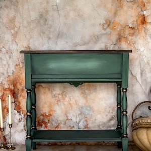 May include: A dark green console table with a dark brown top and a lower shelf. The table has decorative turned legs with gold accents. The background is a textured wall with a mix of brown, orange, and white tones.
