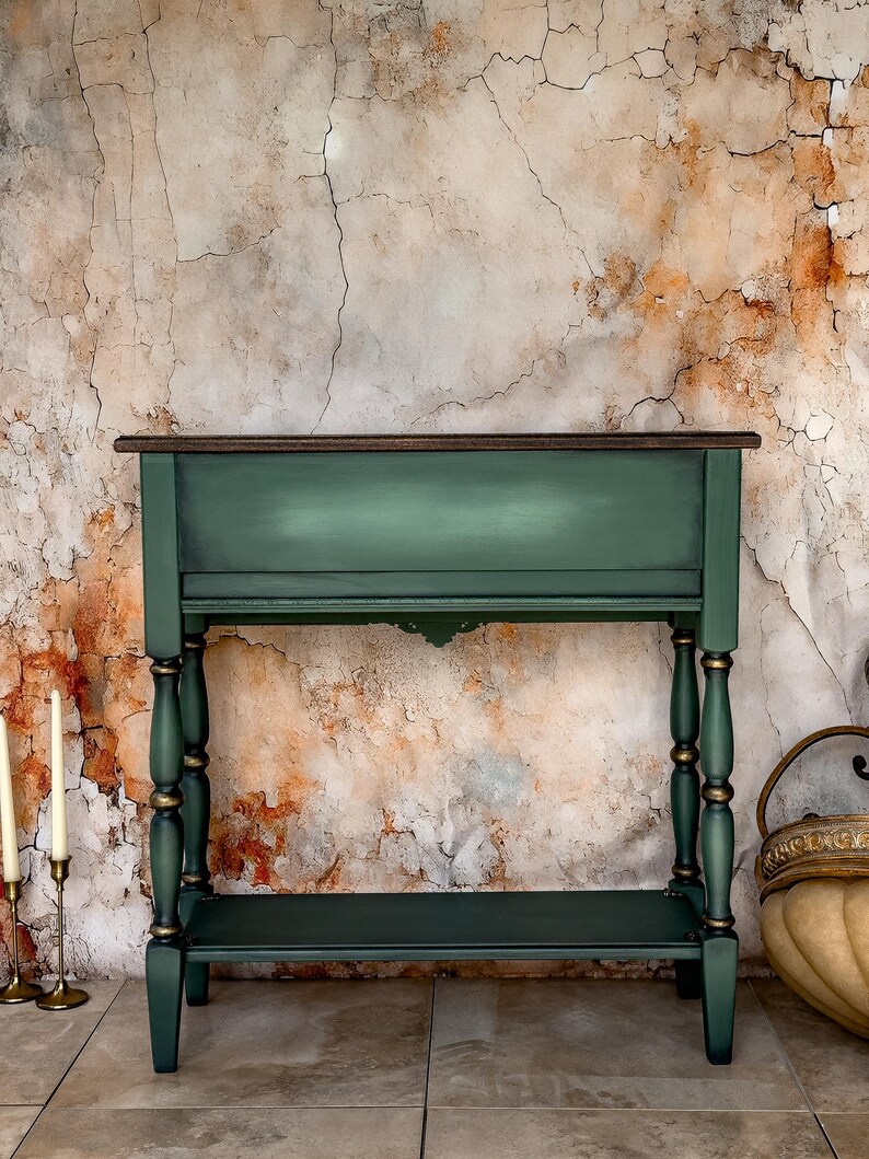 May include: A dark green console table with a dark brown top and a lower shelf. The table has decorative turned legs with gold accents. The background is a textured wall with a mix of brown, orange, and white tones.