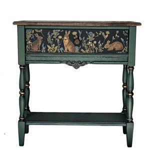 Hand-painted green console table featuring a woodland rabbit design on the front panel, shown isolated on a white background. The piece has turned legs, subtle gold accents, and a lower shelf.