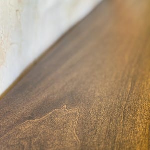 May include: Close-up of a stained wooden surface with visible wood grain. The wood has a warm, brown tone with lighter highlights, suggesting a natural finish. The surface appears smooth and well-maintained.