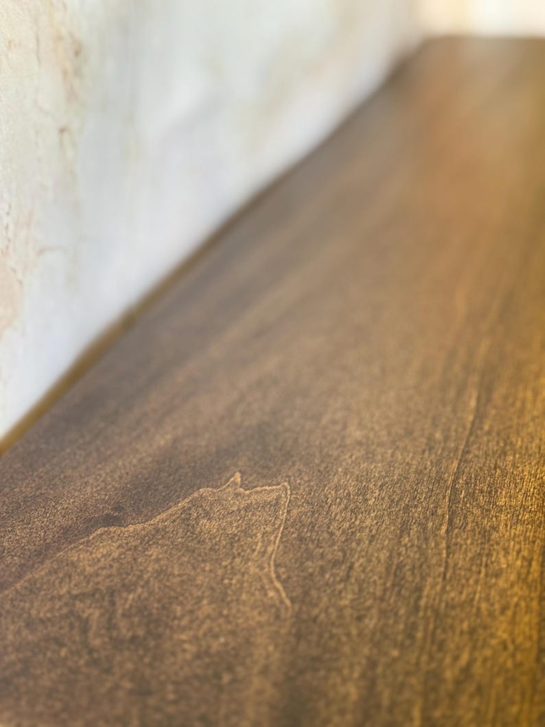 May include: Close-up of a stained wooden surface with visible wood grain. The wood has a warm, brown tone with lighter highlights, suggesting a natural finish. The surface appears smooth and well-maintained.