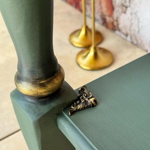 May include: Close-up of a green painted wooden table leg with a gold-painted base. A small, ornate gold-colored metal corner bracket is attached to the table. Two gold candle holders are in the background.