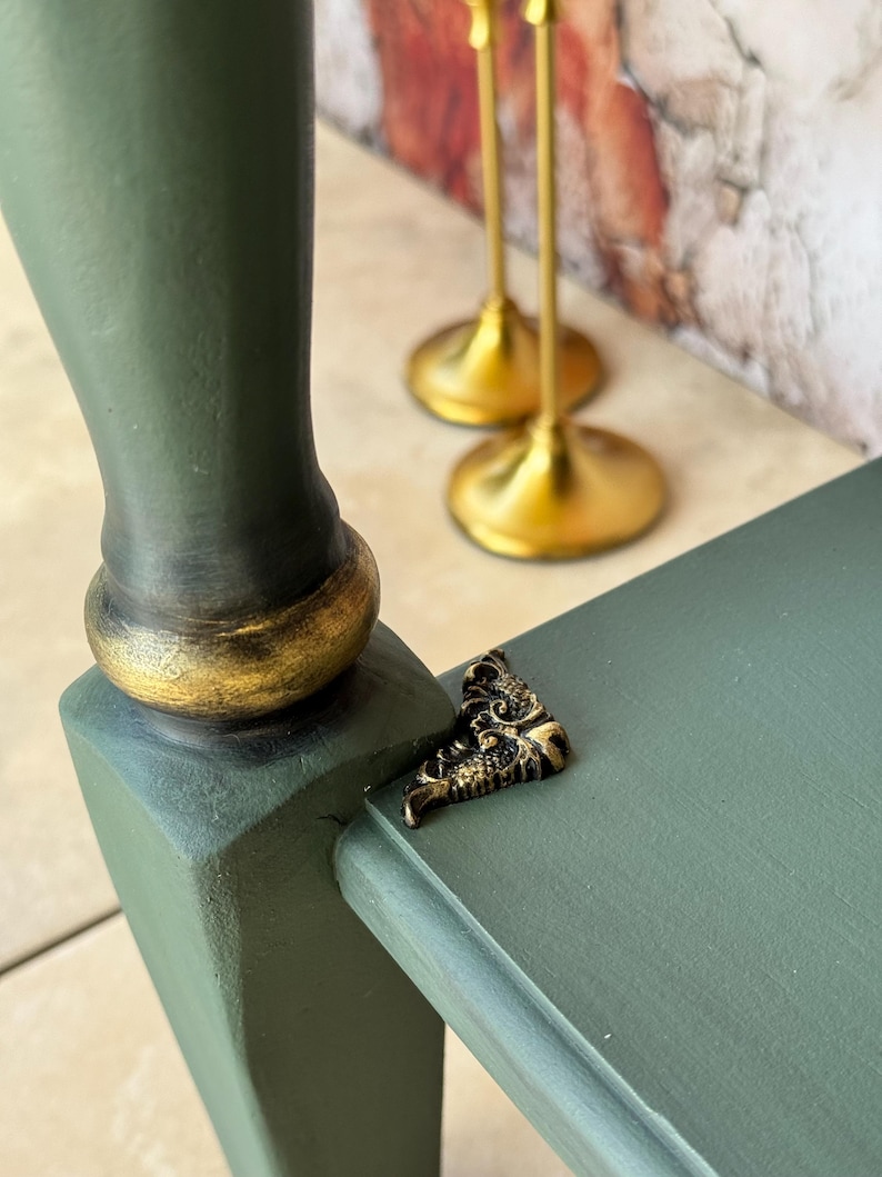 May include: Close-up of a green painted wooden table leg with a gold-painted base. A small, ornate gold-colored metal corner bracket is attached to the table. Two gold candle holders are in the background.