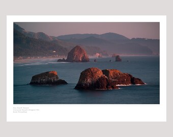 May include: A scenic photograph of a rocky coastline at sunset. The image features several large rock formations jutting out of the water, with a hazy mountain range in the background. The sky is a soft pink and orange, and the water is a deep blue.