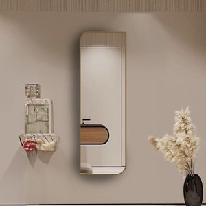 May include: A modern interior scene featuring a tall, rectangular mirror with a gold frame, a unique wall-mounted sculpture, and a black vase with dried flowers. The room has a neutral color palette with a light-colored rug.