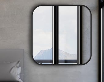 Pode incluir: Um espelho de parede quadrado com cantos arredondados e uma moldura preta fina. O espelho está montado numa parede cinzenta acima de uma cama com roupa de cama cinzenta. A superfície reflete uma moldura de janela e uma paisagem montanhosa.