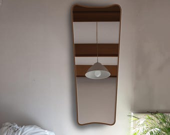 Pode incluir: Um espelho de parede alto e vertical com uma moldura de madeira lisa em tom castanho claro, apresentando uma forma distinta ligeiramente afunilada com bordas curvas. O espelho reflete o ambiente, incluindo uma luminária pendente e vigas.