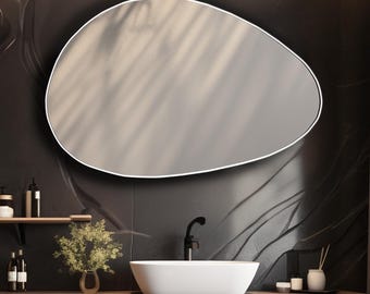 Pode incluir: Um espelho de parede com formato irregular de seixo e uma moldura branca fina, montado sobre um lavatório branco. O lavatório assenta num móvel de madeira escura com grão horizontal, torneira preta e vários acessórios de casa de banho.
