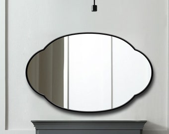 May include: A wall-mounted mirror featuring a black frame with a distinctively curved, scalloped oval shape. The reflective glass surface displays a clear view of the surrounding interior space, including a fireplace and a modern light fixture.