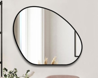 Pode incluir: Um espelho de parede assimétrico com uma moldura preta fina. O espelho apresenta uma forma orgânica irregular que reflete o espaço interior circundante, incluindo uma janela e paredes de cores claras, proporcionando uma superfície refletora.