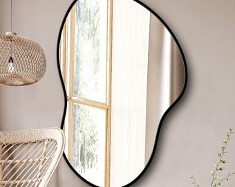 Pode incluir: Um espelho de parede com formato orgânico irregular e uma moldura preta fina e lisa. A superfície reflexiva exibe uma visão clara de uma janela, capturando a luz natural e criando uma aparência brilhante e aberta no espaço.