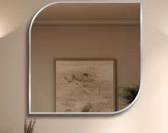 Pode incluir: Um espelho de parede com moldura prateada apresentando um formato assimétrico único com um canto arredondado. A superfície reflexiva mostra o interior de um cômodo com um quadro emoldurado e uma planta, criando um reflexo claro e detalhado.