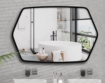 Pode incluir: Um espelho de parede com moldura preta e formato hexagonal arredondado. A superfície refletora exibe o interior de um banheiro com uma banheira branca independente, um box de chuveiro e uma janela com persianas horizontais.