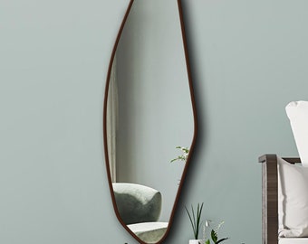 Pode incluir: Um espelho de parede alto com formato orgânico irregular e uma moldura fina castanho-escura. O espelho apresenta uma silhueta assimétrica e lisa que se estreita no topo e alarga na base, refletindo o interior de uma sala com cadeira e plantas.