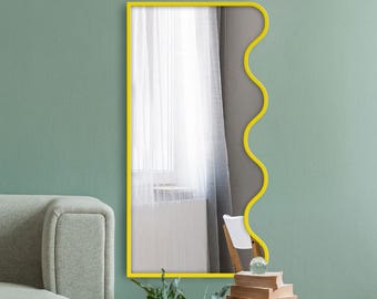 Pode incluir: Um espelho de parede retangular com uma moldura amarela brilhante apresentando uma borda ondulada e recortada distinta no lado direito. O espelho reflete uma janela com cortinas brancas e parte de uma cadeira de madeira, exibindo um design moderno.