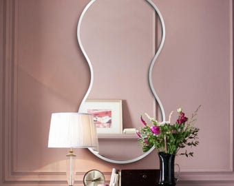 Pode incluir: Um espelho de parede com uma moldura metálica prateada distinta em forma de amendoim alongado. O espelho apresenta um contorno curvo suave e contínuo que cria uma silhueta única, refletindo a sala e os objetos posicionados à sua frente.