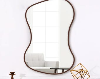 Pode incluir: Um espelho de parede com uma forma irregular, orgânica e ameboide, com uma moldura fina castanho-escura. A superfície refletora mostra uma visão clara do interior de uma divisão, incluindo uma cadeira branca e uma janela com grelha.