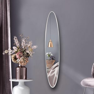 Puede incluir: Un espejo de cuerpo entero de forma ovalada blanca con un marco blanco fino. El espejo está colgado en una pared gris y refleja una habitación con una lámpara, una mesa auxiliar y un jarrón de flores.