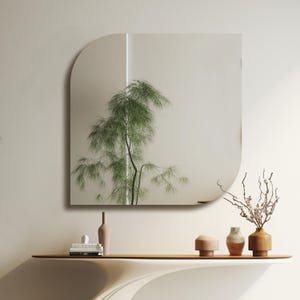 May include: A white wall with a curved, square mirror hanging on it. The mirror reflects a green plant and a white wall. A white shelf with three brown ceramic vases and a book sits below the mirror.