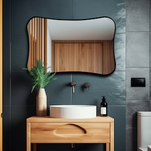 Può includere: Uno specchio con cornice nera dalla forma unica è appeso sopra un mobiletto in legno con un lavandino bianco. Una pianta verde in un vaso beige e un dosatore di sapone nero sono sul mobiletto. Il bagno ha piastrelle verde scuro e dettagli in legno.