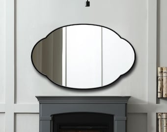 May include: An oval-shaped wall mirror featuring a thin black frame with a scalloped edge design. The mirror is mounted on a light gray wall above a dark gray fireplace mantel, reflecting the interior space and a modern light fixture above.