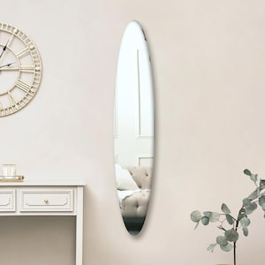 May include: A full-length oval mirror with a silver frame. The mirror is hanging on a white wall and reflects a room with a white sofa.