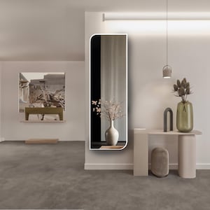 May include: A white framed full-length mirror with rounded edges, reflecting a room with a vase of white flowers and a picture on the wall. The mirror is mounted on a white wall.