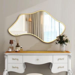 May include: An irregularly shaped gold-framed mirror hangs above a white vanity with gold accents. The mirror reflects a room with curtains and a hanging light. A vase of flowers and decorative items sit on the vanity.