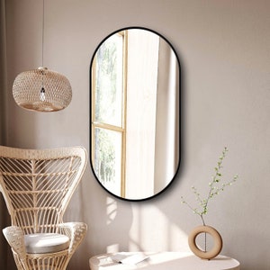 Puede incluir: Un espejo ovalado negro con un marco negro delgado cuelga de una pared beige claro. El espejo refleja una ventana con cortinas blancas y una vista de &aacute;rboles verdes en el exterior.