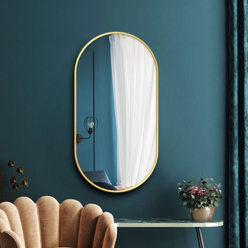 Puede incluir: Un espejo ovalado dorado con un borde biselado cuelga de una pared azul verdosa. El espejo refleja una cortina blanca y una l&aacute;mpara.