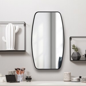 May include: A modern vanity setup featuring a large, oval mirror with a black frame. Two black metal cube shelves hold a white cactus sculpture and decorative items. Makeup brushes, containers, and a candle sit on the white tabletop.