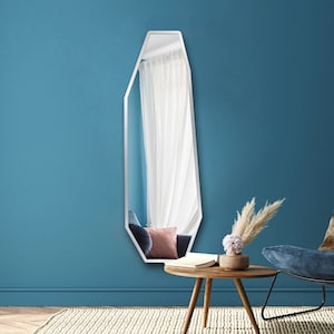 Puede incluir: Un espejo alto con marco blanco y forma geométrica irregular. Refleja una habitación con un sillón azul, un cojín rosa y cortinas blancas. Está montado en una pared azul turquesa, con una pequeña mesa de madera y objetos decorativos.
