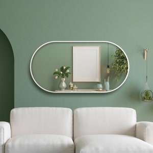 Puede incluir: Un espejo blanco de forma ovalada con un estante, que presenta un cuadro enmarcado, un jarr&oacute;n con flores rosas, dos velas y un peque&ntilde;o jarr&oacute;n azul. El espejo est&aacute; colgado en una pared verde salvia sobre un sof&aacute; blanco.