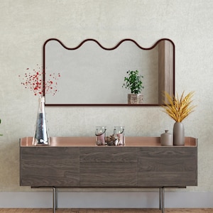 Könnte beinhalten: Ein modernes Wohnzimmer mit einer dunklen Holzkommode mit kupferfarbener Oberseite. Darüber hängt ein wellenförmiger, braun gerahmter Spiegel. Dekorative Vasen und Kerzen schmücken die Kommode, zusammen mit einer kleinen Pflanze und einer Vase mit Trockenblumen.