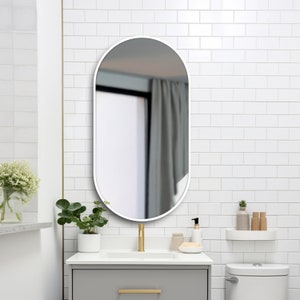 Puede incluir: Un espejo de ba&ntilde;o ovalado blanco con marco blanco cuelga de una pared de azulejos blancos sobre un tocador de ba&ntilde;o blanco con un grifo dorado.