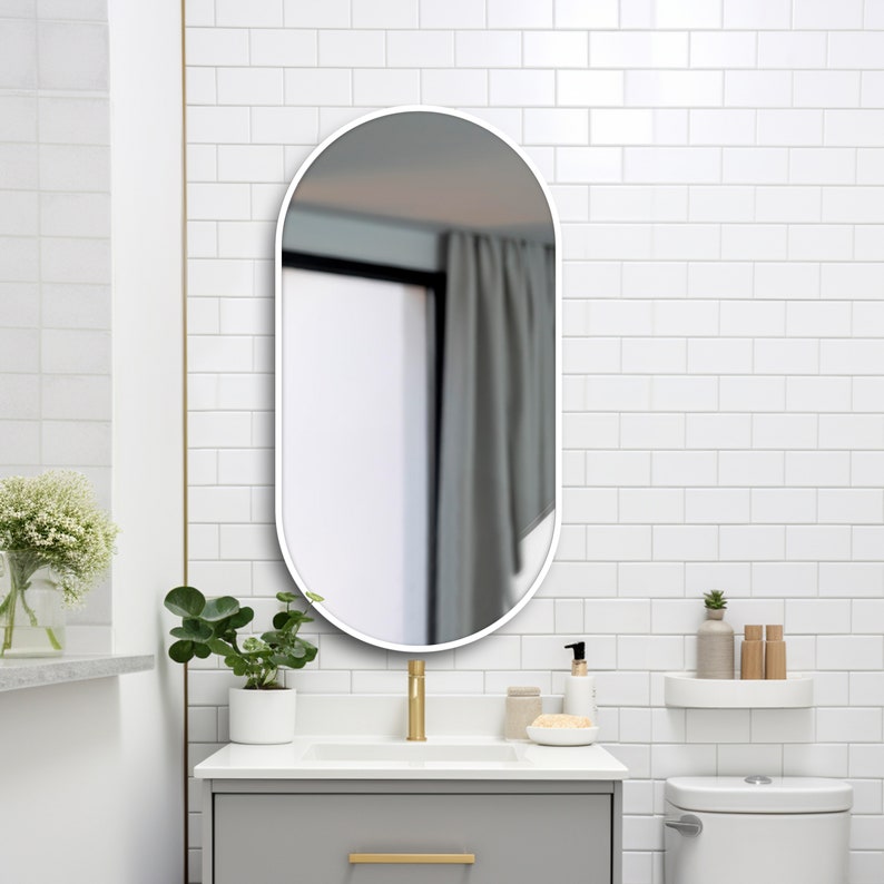 Puede incluir: Un espejo de ba&ntilde;o ovalado blanco con marco blanco cuelga de una pared de azulejos blancos sobre un tocador de ba&ntilde;o blanco con un grifo dorado.