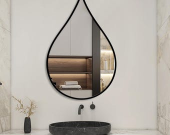 Pode incluir: Um espelho de parede em forma de gota com uma moldura preta fina. A superfície refletora mostra uma visão clara do interior de uma sala com prateleiras de madeira e armários brancos. O espelho está montado numa parede sobre um lavatório.