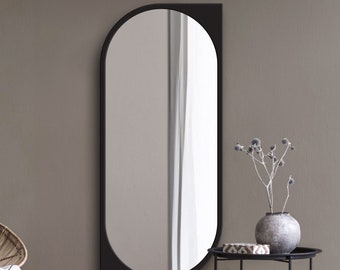Pode incluir: Um espelho de parede com moldura preta de formato assimétrico único, combinando uma borda vertical reta no lado direito com um topo arredondado e um lado esquerdo curvo, refletindo um espaço interior iluminado com uma cortina branca.