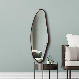May include: A wall-mounted mirror with an irregular, organic, pebble-like shape and a thin, dark brown frame. The reflective surface shows a clear view of the interior space, capturing the light and depth of the room within its unique, asymmetrical border.
